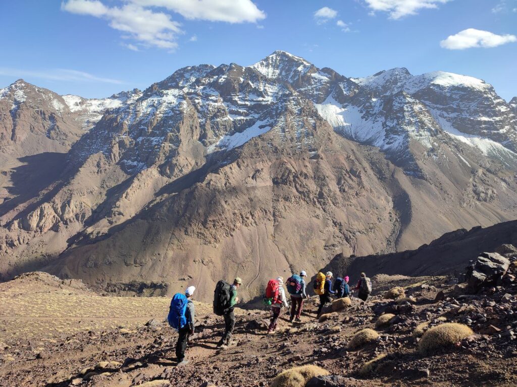 Trekkers descending a rocky trail below the snow-capped Toubkal massif in Morocco's High Atlas